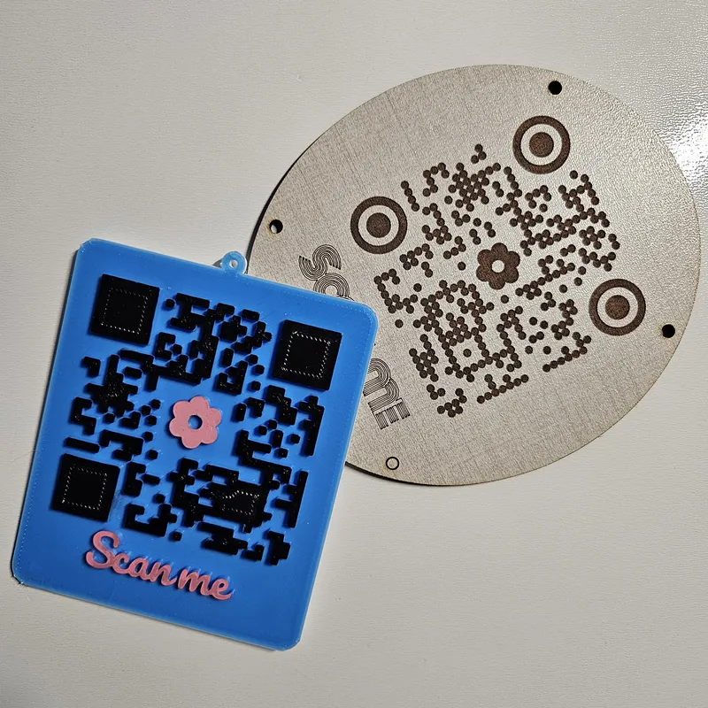 QR code signs: blue PLA 3D-printed plaque with Square module style, pink flower logo and Scan Me label next to a laser-engraved TruFlat Linen plywood plaque with dot pattern styling